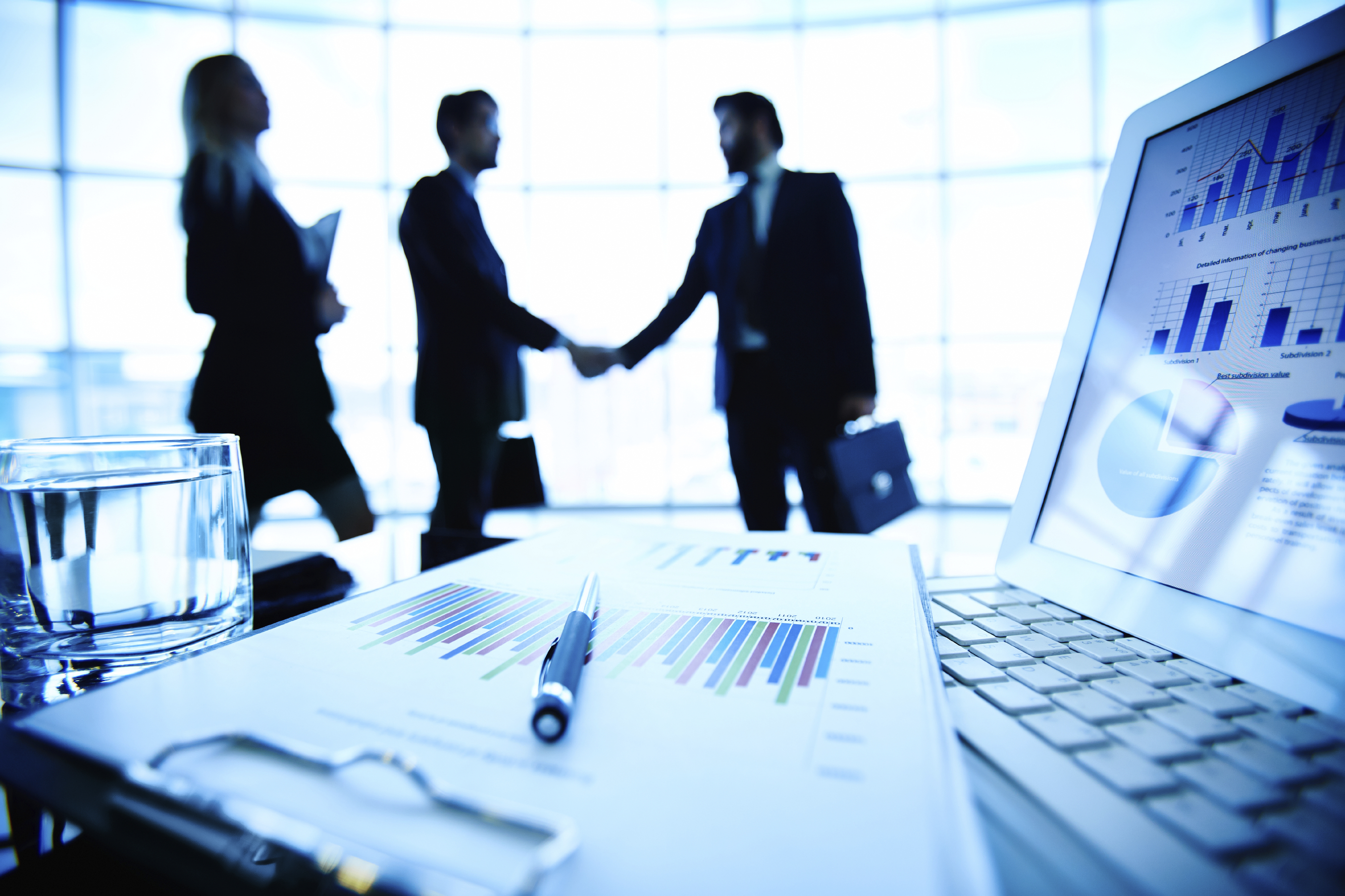 Laptop, financial document with pen and glass of water at workplace on background of three business partners making a deal